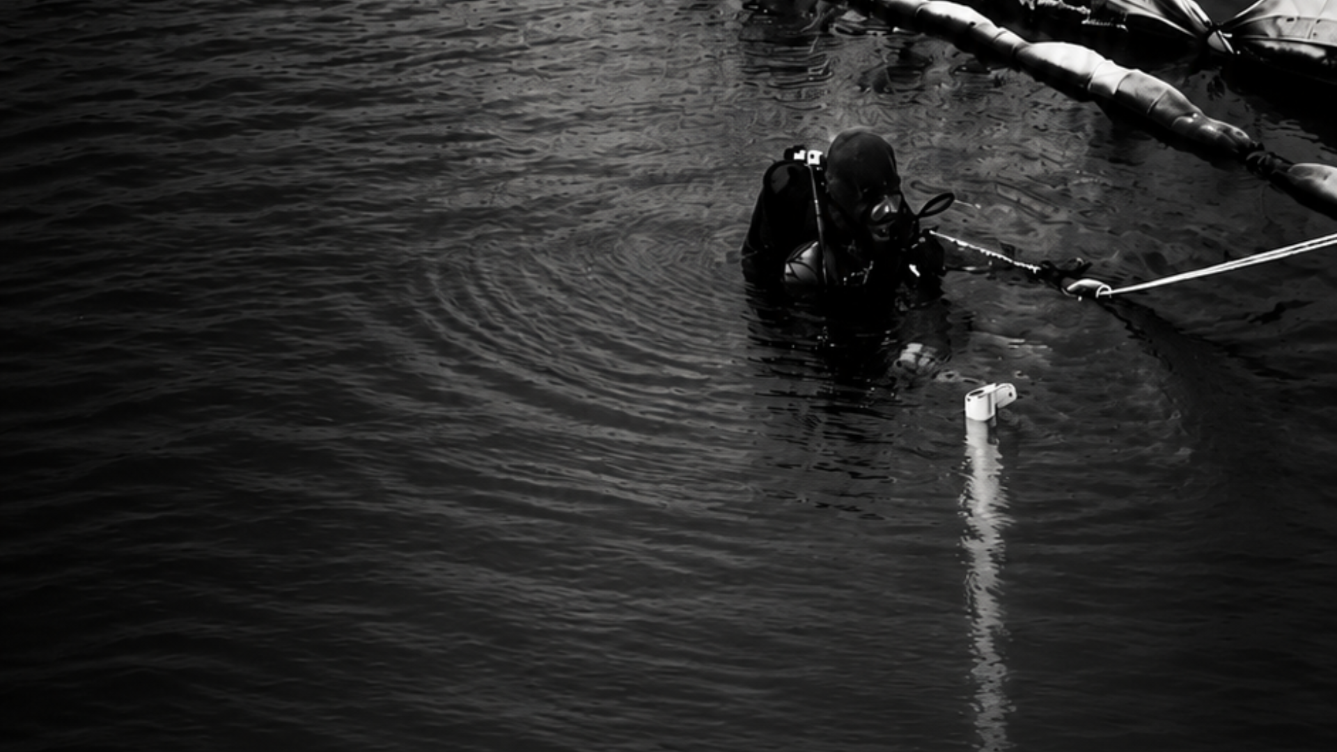 Black-and-white photograph of divers working in dark water beside submerged equipment.